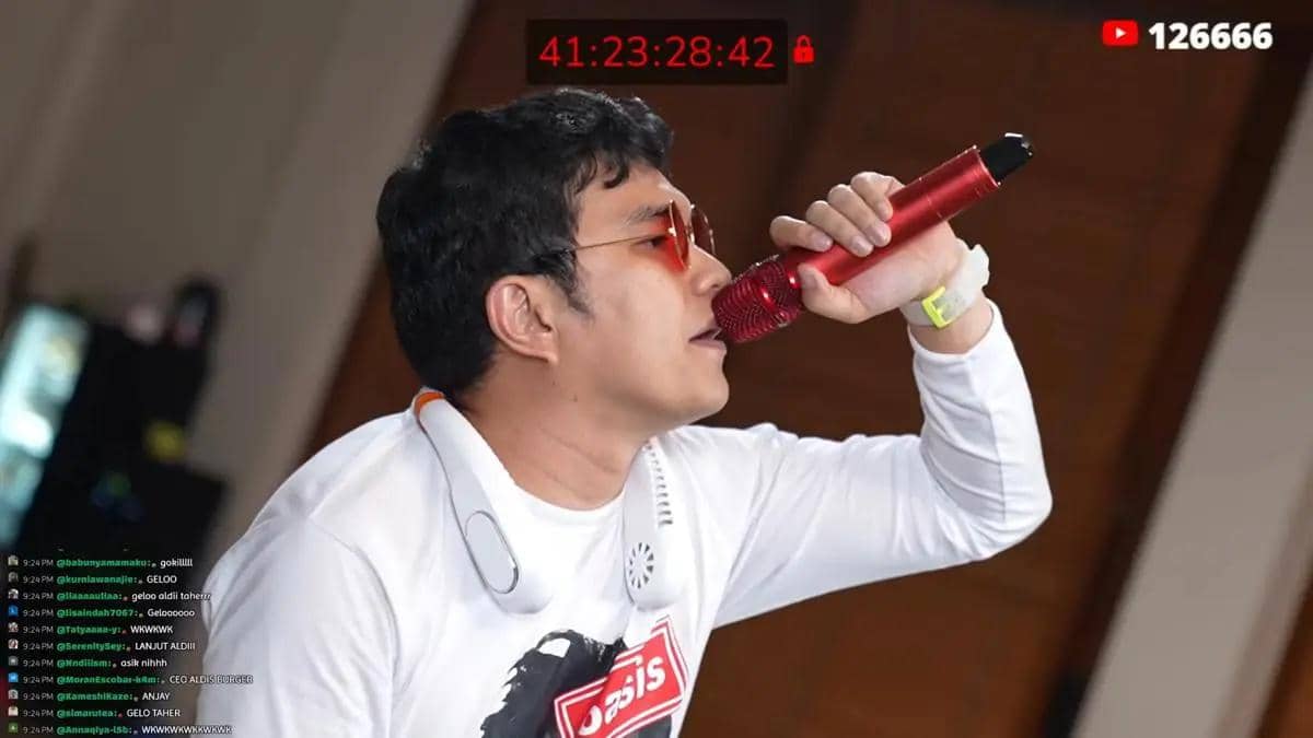Seorang pria mengenakan kacamata hitam dan kaus putih sedang bernyanyi menggunakan mikrofon merah di acara Marapthon Season 3.