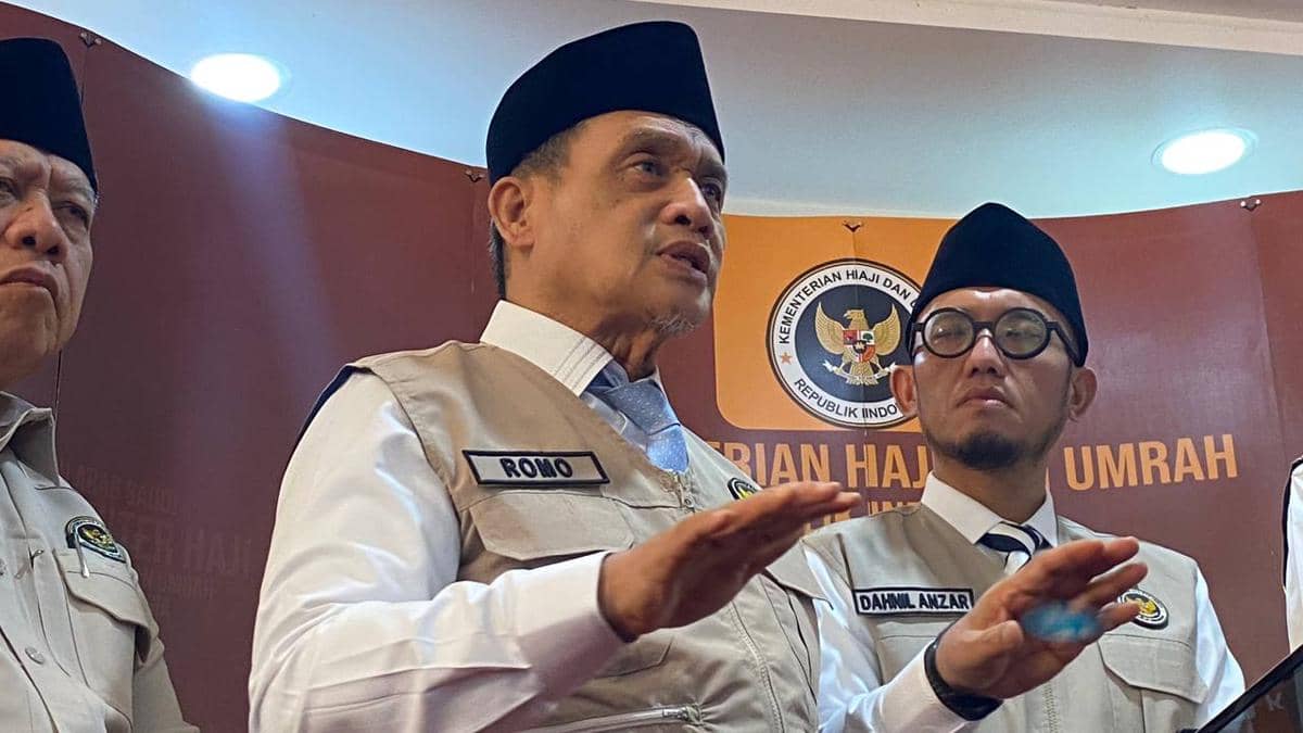 Seorang pejabat Kementerian Agama mengenakan rompi bertuliskan nama dan peci hitam berbicara di depan latar bertema Kementerian Haji dan Umrah.