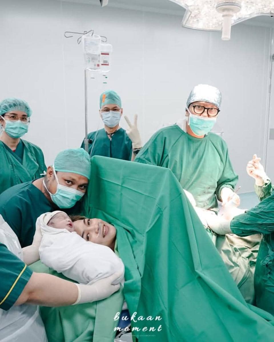 Seorang ibu tersenyum setelah melahirkan di ruang operasi, didampingi tim medis yang mengenakan pakaian hijau dan masker bedah.