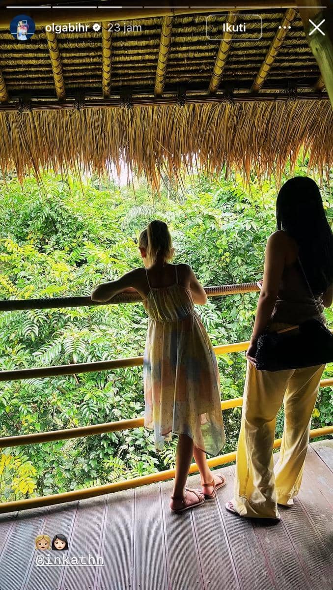 Dua perempuan berdiri di balkon bambu beratap jerami menghadap pemandangan hijau pepohonan tropis di Bali.