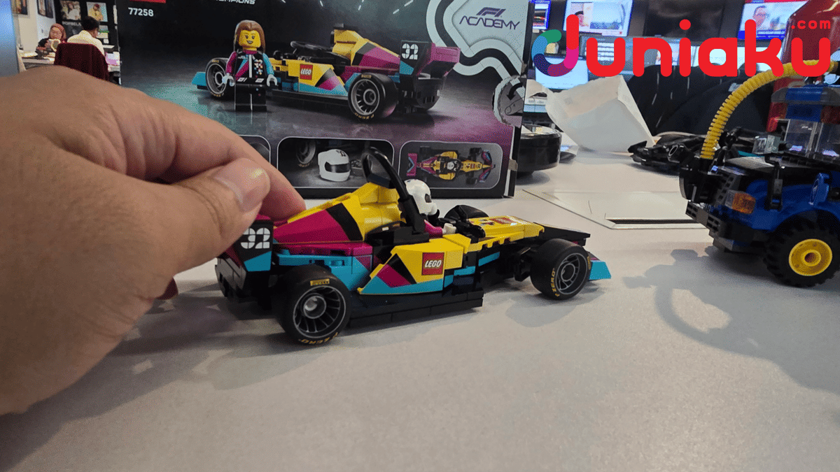 Tangan seseorang memegang mobil balap LEGO Speed Champions F1 Academy berwarna kuning, merah muda, dan biru di atas meja.