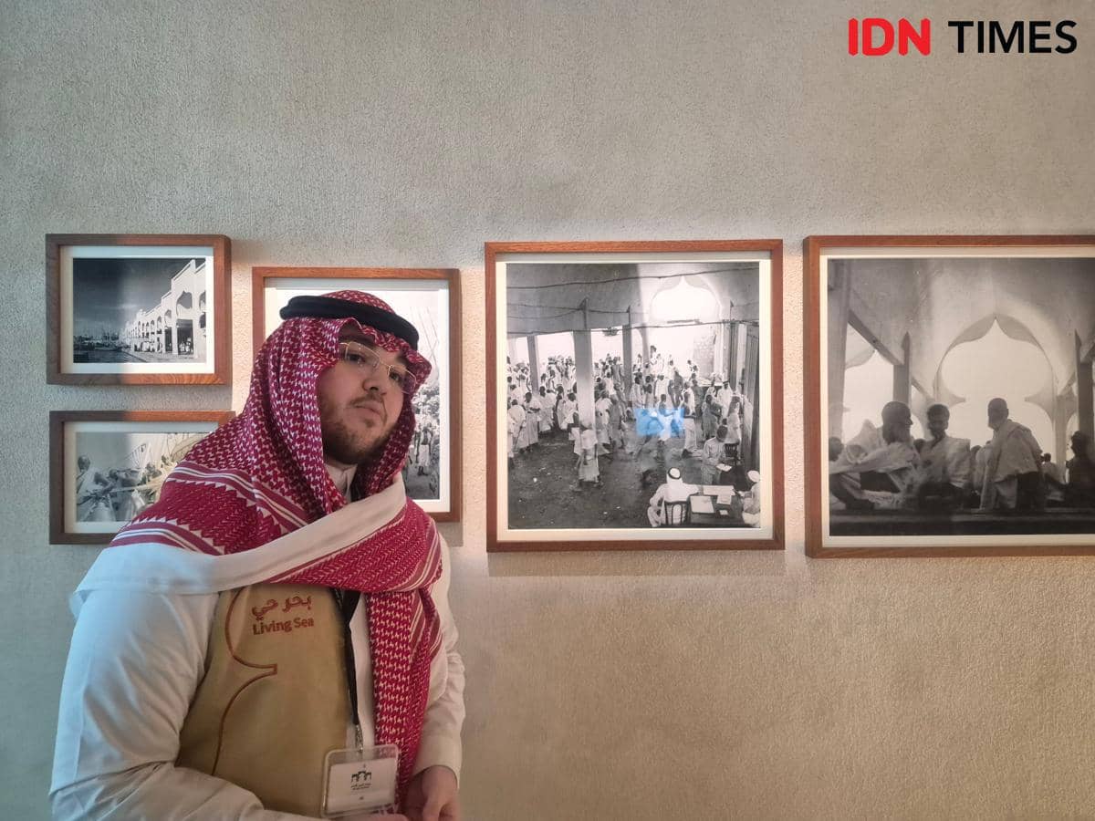 Seorang pria berpakaian tradisional Arab berdiri di depan dinding yang dipenuhi foto hitam putih tentang sejarah kedatangan jemaah haji di Jeddah.
