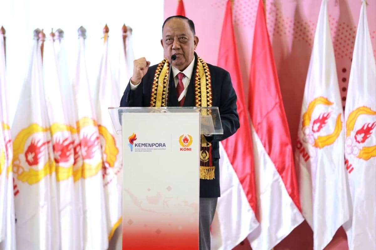 Seorang pria berpidato di podium berlogo Kemenpora dan KONI dengan latar bendera Indonesia serta bendera KONI di acara Rakernas dan Musornaslub 2026.