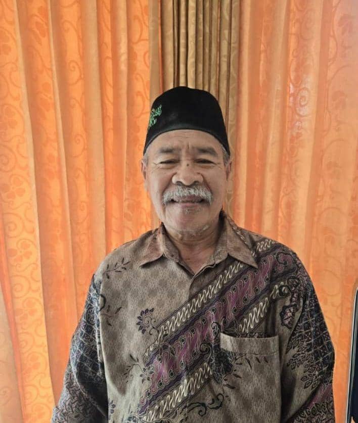 Seorang pria lanjut usia mengenakan peci hitam dan batik cokelat berdiri tersenyum di depan tirai oranye bermotif.