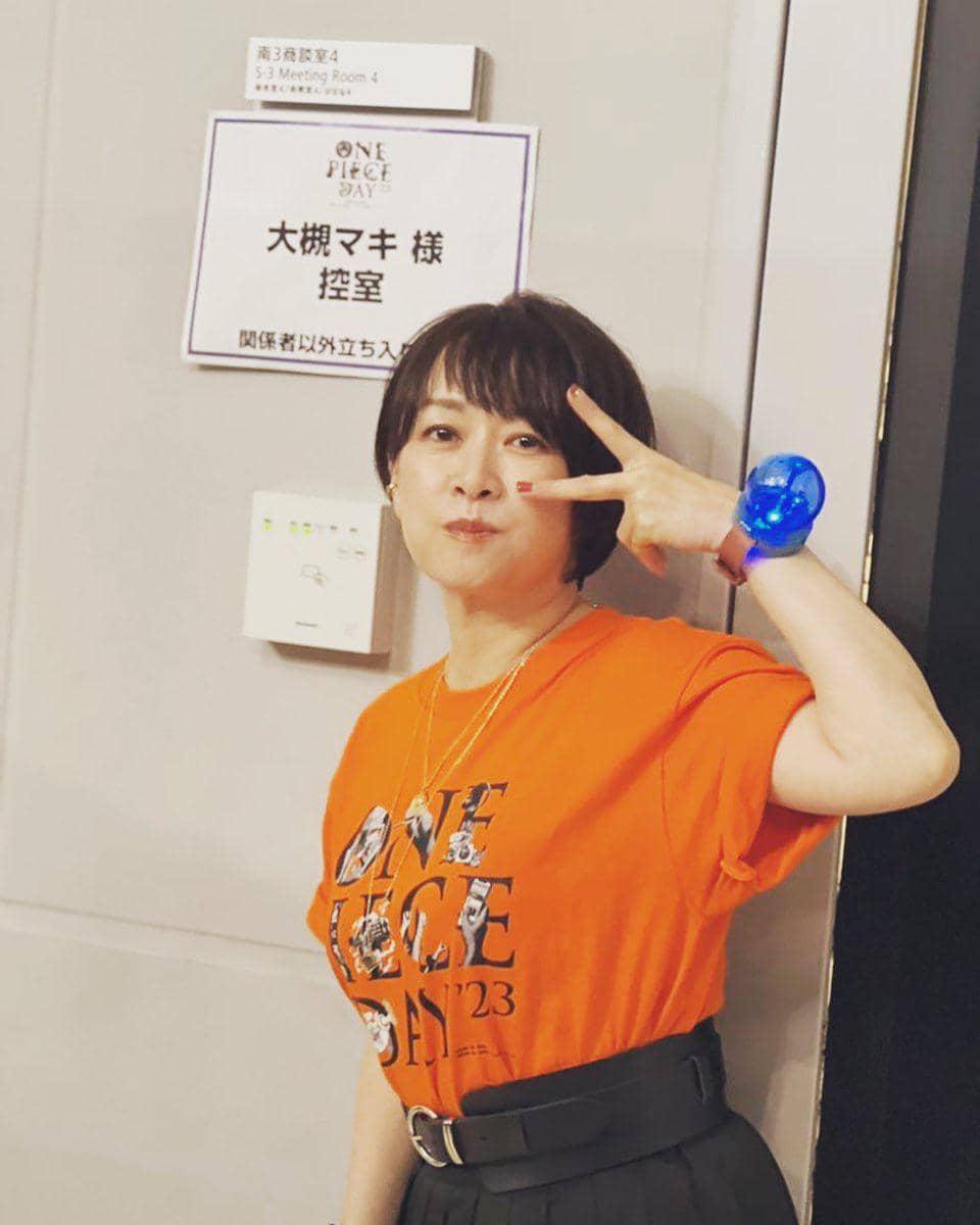 Seorang wanita berpose dengan tanda tangan damai di depan pintu ruang ganti bertuliskan One Piece Day, mengenakan kaus oranye bertema acara tersebut.