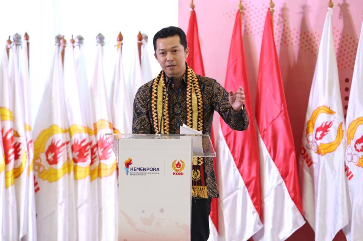 Seorang pria berbatik memberikan sambutan di podium berlogo Kemenpora dan KONI dengan latar bendera Indonesia dan bendera KONI.