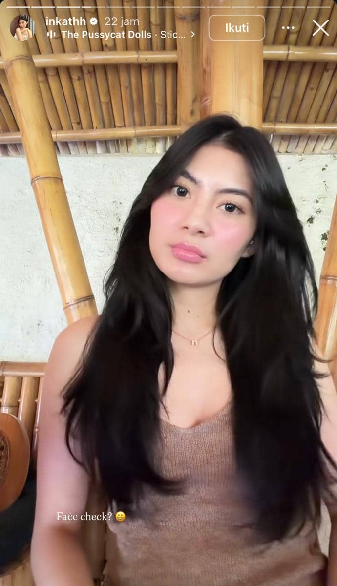 Seorang perempuan berambut panjang mengenakan tank top cokelat duduk di depan dinding dan tiang bambu dengan ekspresi santai.