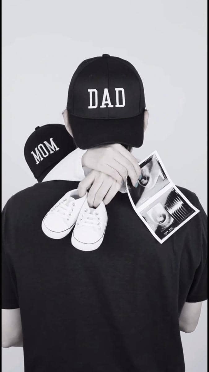 Sepasang calon orang tua mengenakan topi bertuliskan 'MOM' dan 'DAD', memegang sepatu bayi putih dan hasil USG di latar abu-abu.