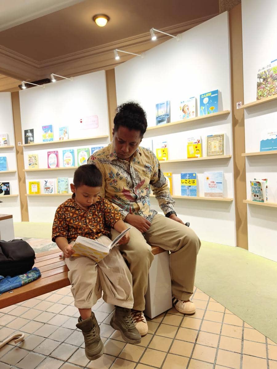 Seorang pria dan anak kecil mengenakan batik duduk di bangku kayu sambil membaca buku di ruang baca dengan rak berisi buku anak-anak.