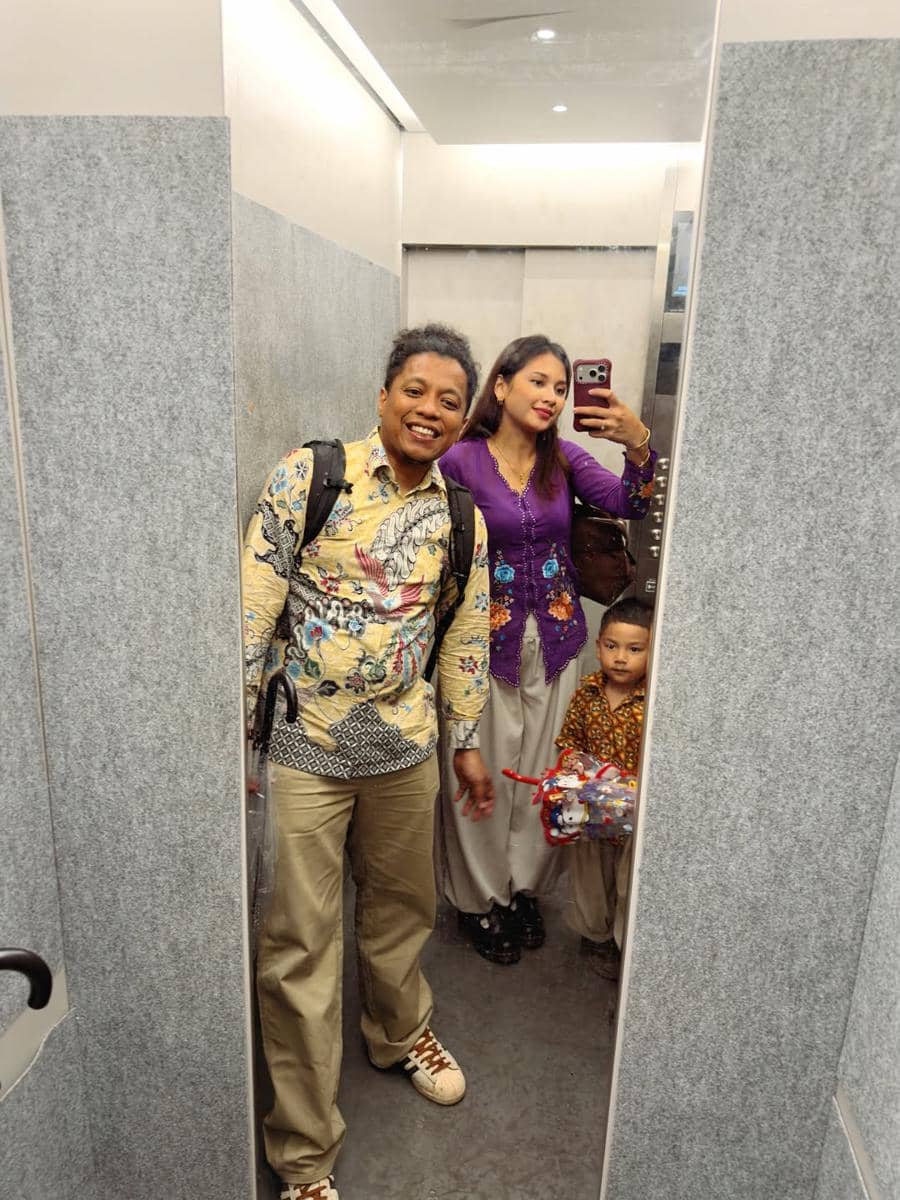 Sepasang orang dewasa dan seorang anak berpose di depan cermin lift mengenakan batik dan kebaya dengan latar interior abu-abu modern.