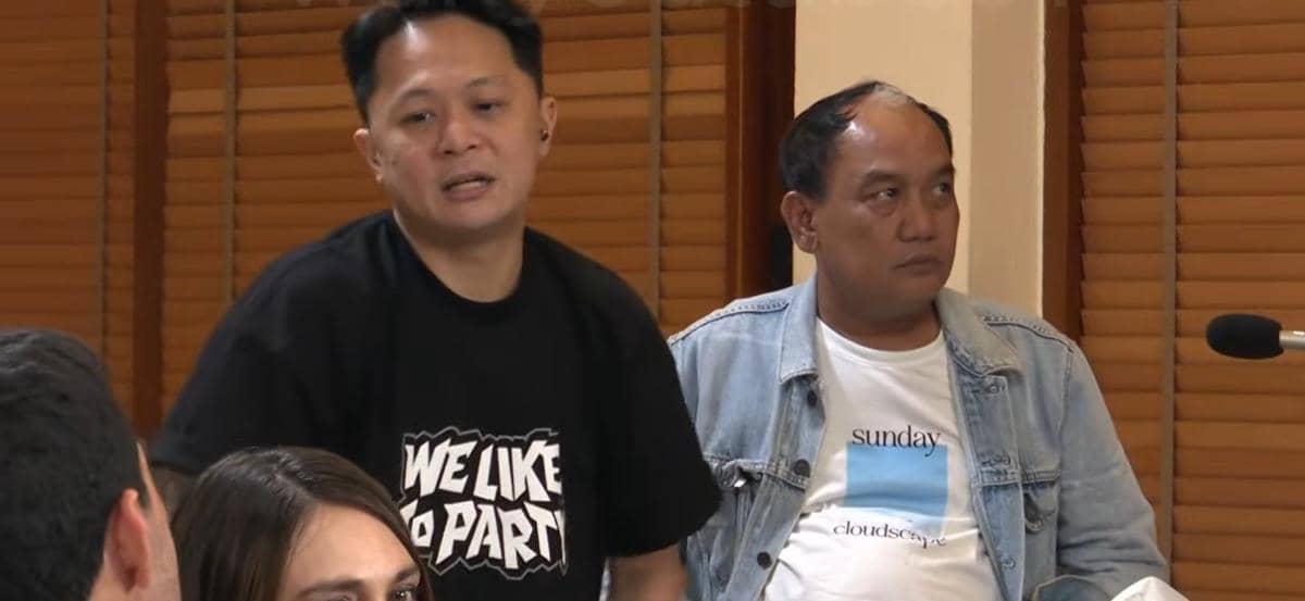 Dua pria duduk berdiskusi di dalam ruangan dengan latar jendela kayu, salah satunya mengenakan kaus hitam dan lainnya jaket denim.