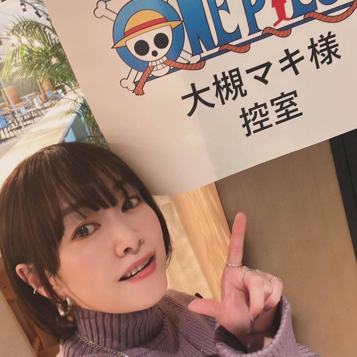 Seorang wanita berpose sambil menunjuk papan bertuliskan logo One Piece dengan huruf Jepang di sebuah ruangan bercahaya hangat.