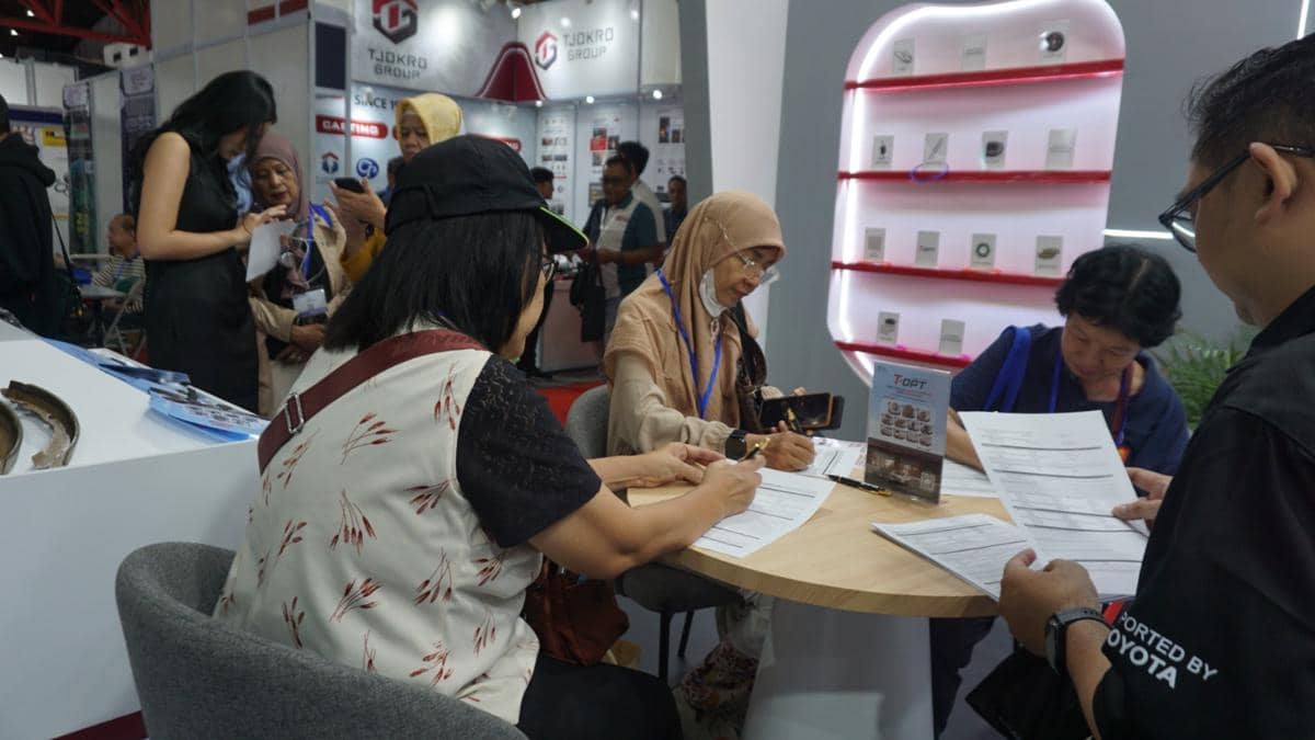 Beberapa orang duduk di meja pameran T-OPT membahas dokumen terkait suku cadang mobil Toyota di sebuah acara otomotif.