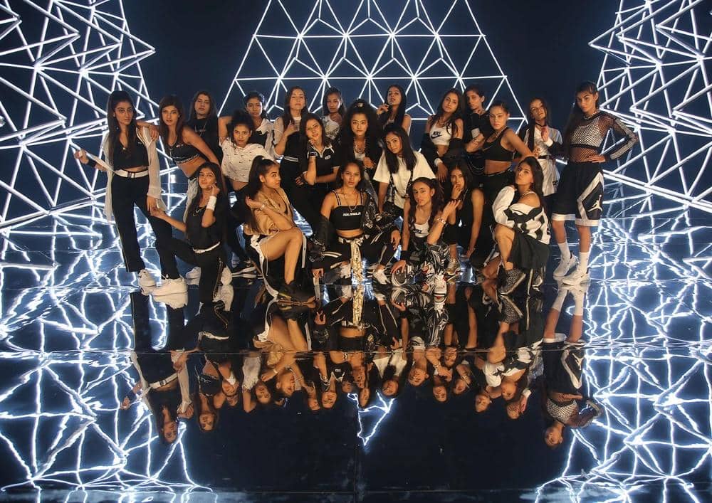 Grup perempuan berpose bersama di atas lantai reflektif dengan latar belakang pola cahaya geometris berwarna putih yang futuristik.
