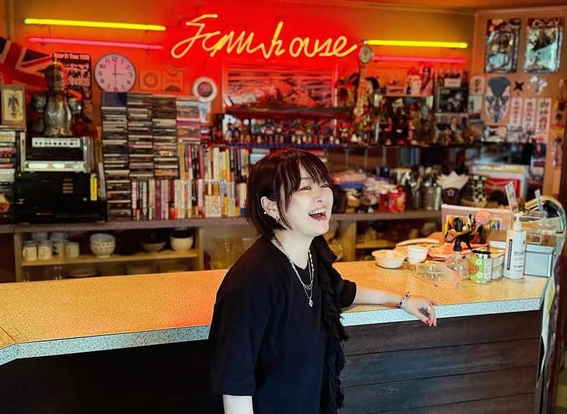 Seorang perempuan berambut pendek mengenakan pakaian hitam tersenyum di depan meja bar dengan latar interior kafe penuh dekorasi warna-warni.