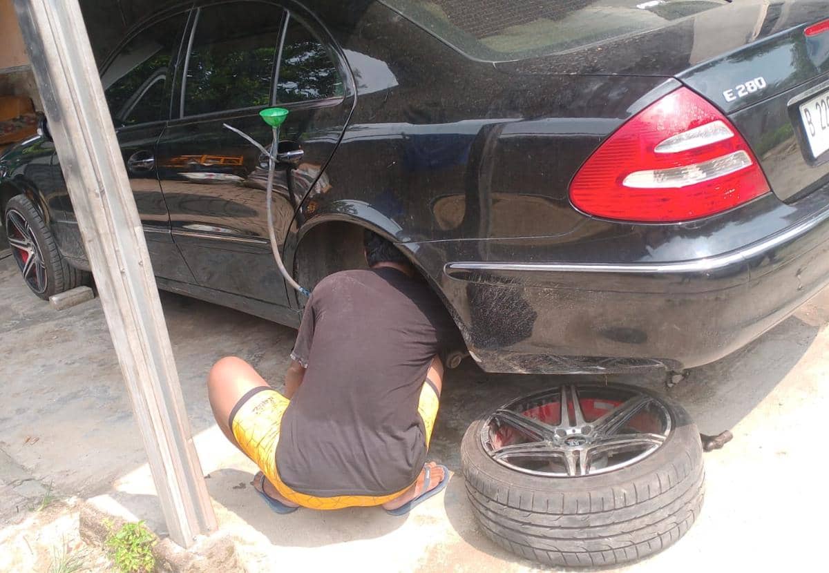 Seorang mekanik memperbaiki mobil sedan hitam di bengkel dengan roda belakang dilepas dan peralatan servis di sekitarnya.