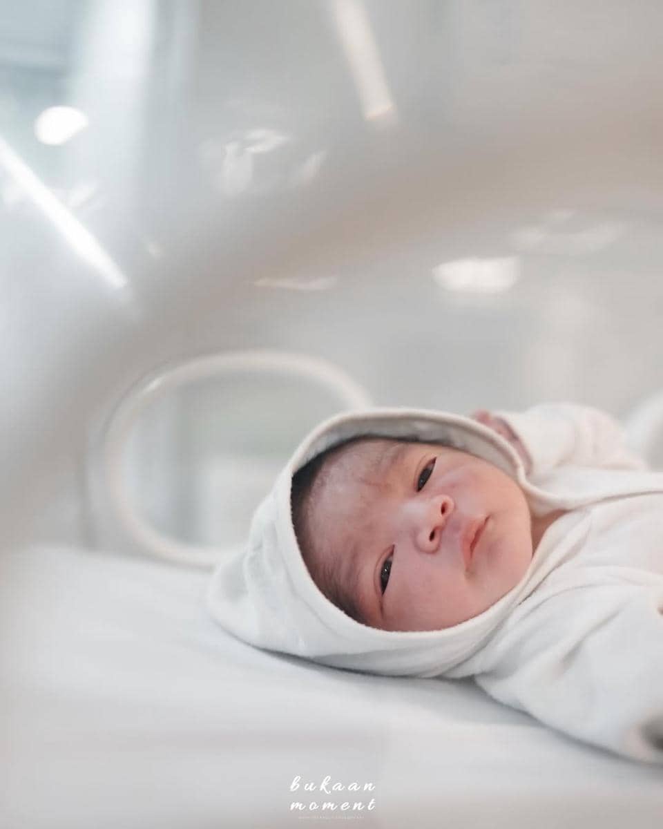 Bayi baru lahir mengenakan pakaian putih dengan tudung, berbaring di tempat tidur rumah sakit dengan pencahayaan lembut.