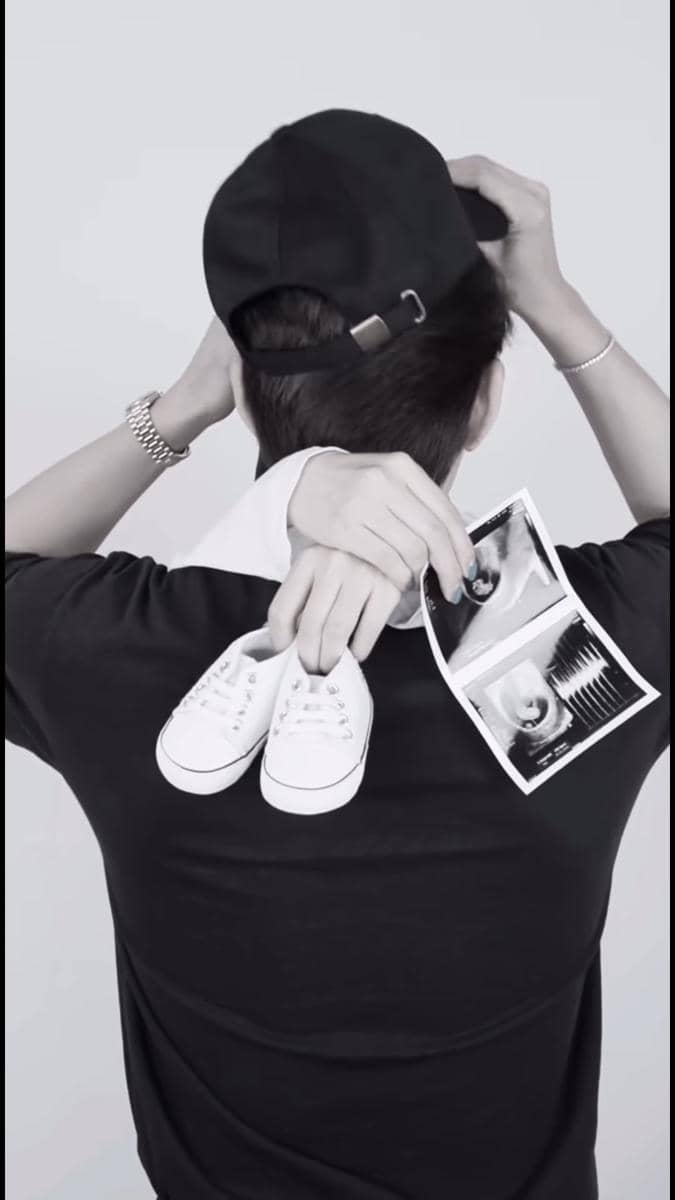 Seorang pria mengenakan topi hitam membelakangi kamera sambil memegang sepatu bayi putih dan hasil USG di tangannya.