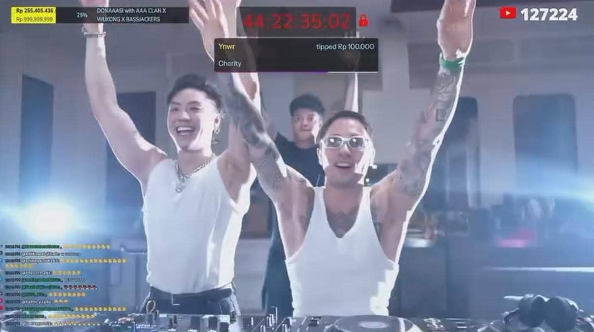 Dua DJ mengenakan kaus tanpa lengan putih mengangkat tangan sambil tersenyum di atas panggung dengan pencahayaan terang dan peralatan DJ.