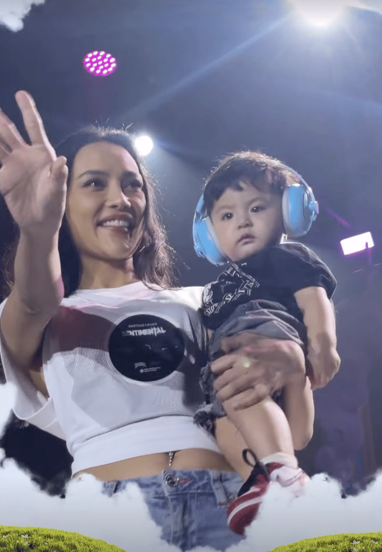 Seorang perempuan tersenyum sambil melambaikan tangan dan menggendong anak kecil yang memakai pelindung telinga biru di acara Marapthon FestivAAAl.