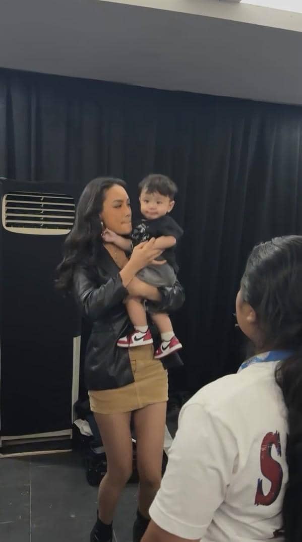 Seorang wanita mengenakan jaket hitam dan rok cokelat muda menggendong anak kecil di ruangan dengan tirai hitam di belakangnya.