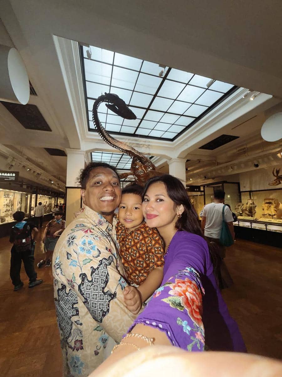 Sebuah keluarga berfoto bersama di museum dengan latar fosil dinosaurus, mengenakan batik dan kebaya berwarna cerah.
