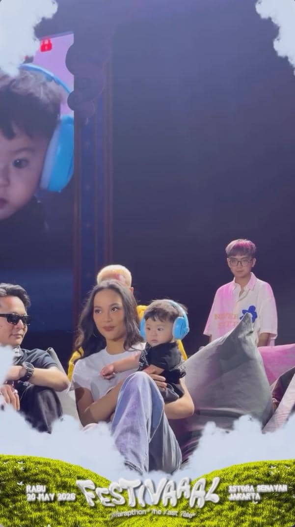 Seorang wanita duduk di sofa sambil menggendong bayi yang mengenakan pelindung telinga biru di acara Festivaaal Marapthon: The Last Tale di Istora Senayan.
