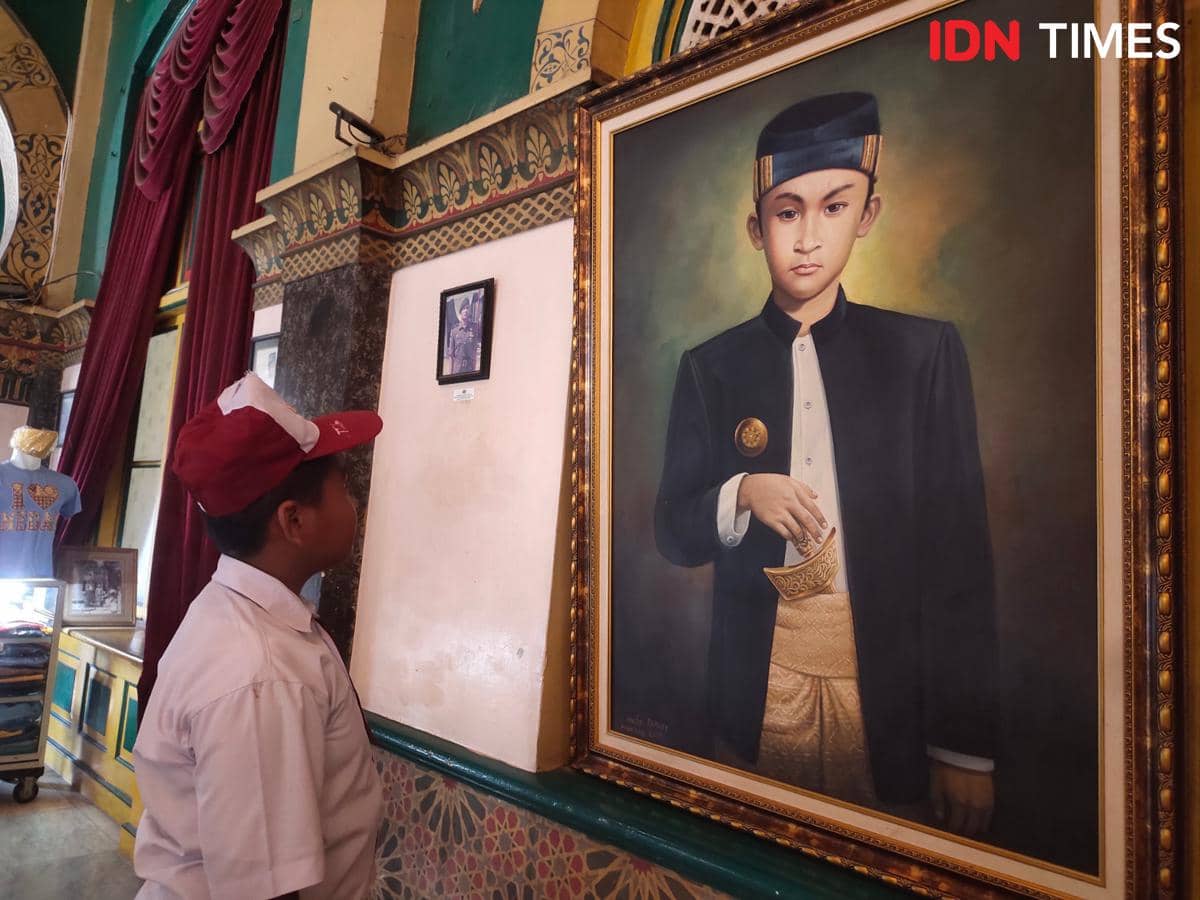 Seorang anak berseragam sekolah dasar melihat lukisan seorang sultan muda berpakaian adat Melayu di dalam ruangan berarsitektur klasik.