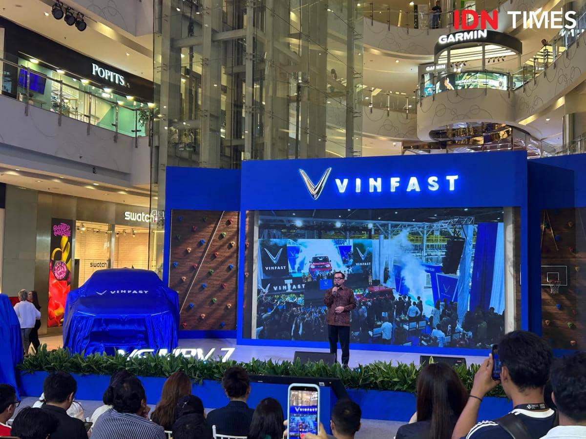 Acara peluncuran mobil VinFast di pusat perbelanjaan dengan panggung biru, layar besar, dan mobil tertutup kain biru di depan penonton.