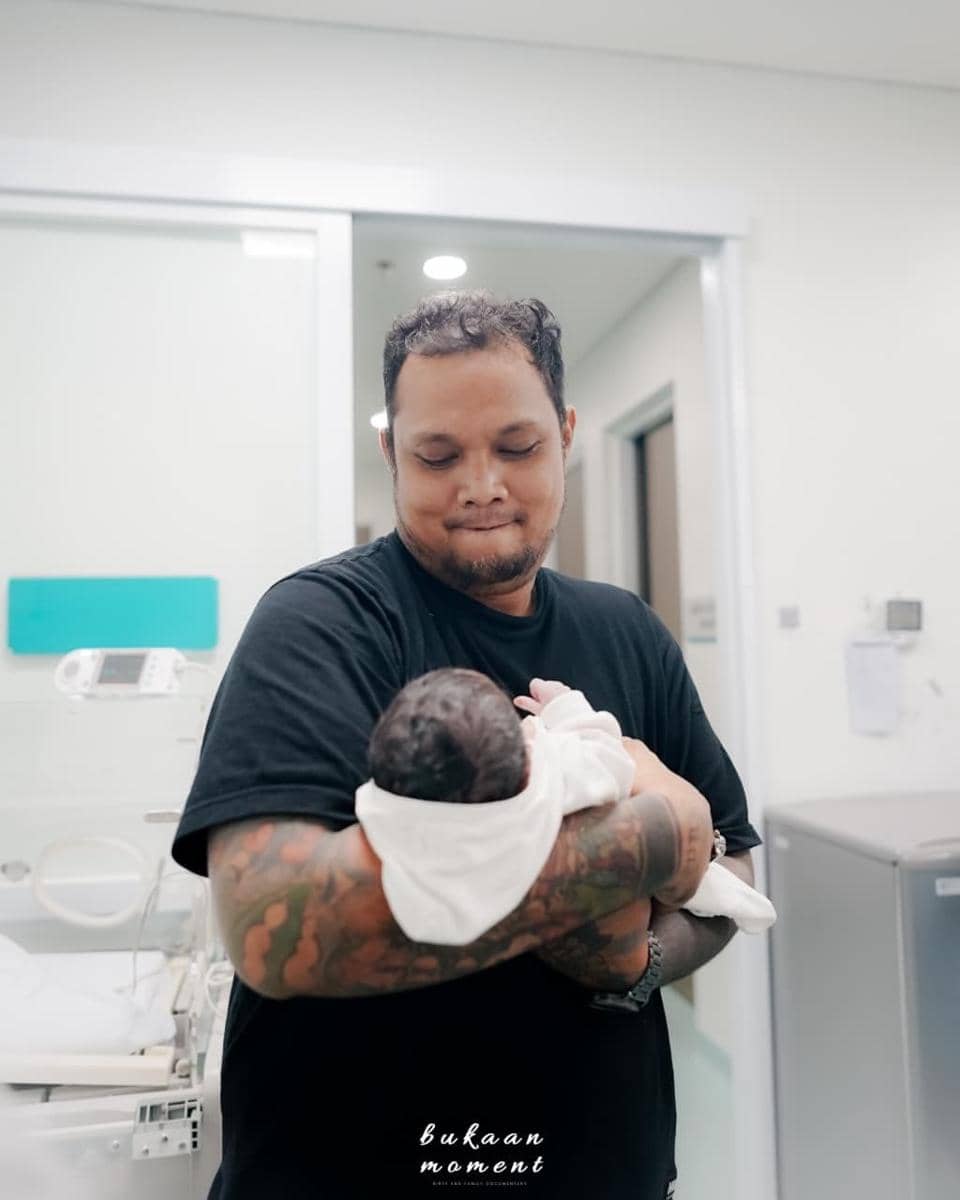 Seorang pria bertato mengenakan kaus hitam menggendong bayi yang baru lahir di ruang rumah sakit dengan ekspresi lembut dan bahagia.