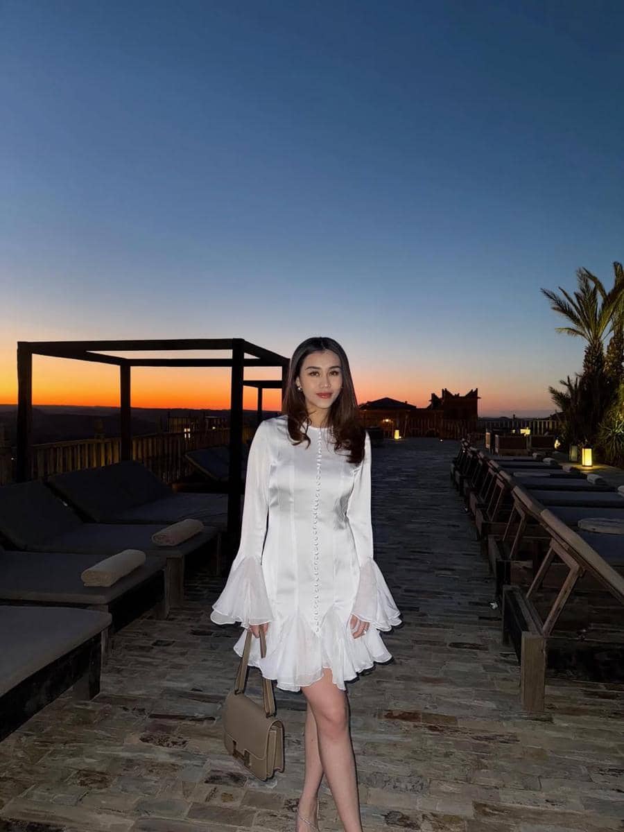 Seorang wanita mengenakan gaun putih elegan dengan tas krem berdiri di area outdoor saat matahari terbenam dengan latar langit senja.