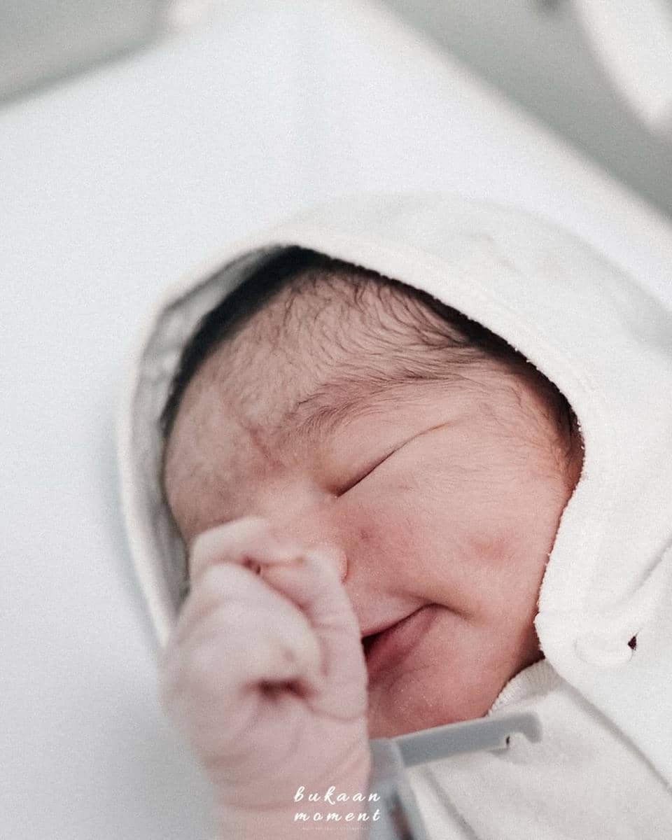 Bayi baru lahir tersenyum dengan mata tertutup, mengenakan selimut putih dan topi bayi, tampak tenang dan hangat.