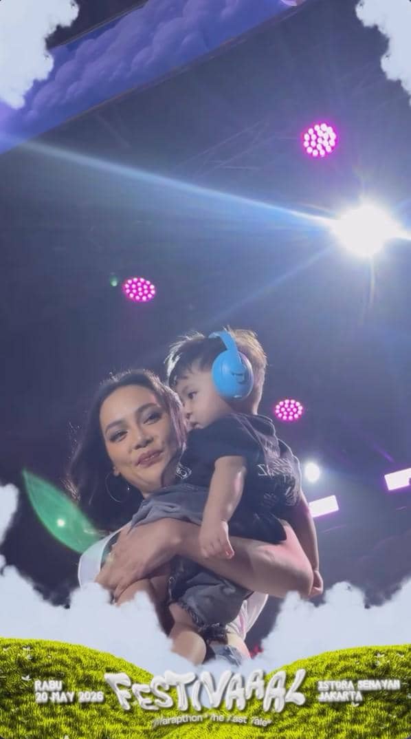 Seorang wanita tersenyum sambil menggendong anak kecil yang memakai pelindung telinga biru di panggung acara Festivaaal Marapthon.