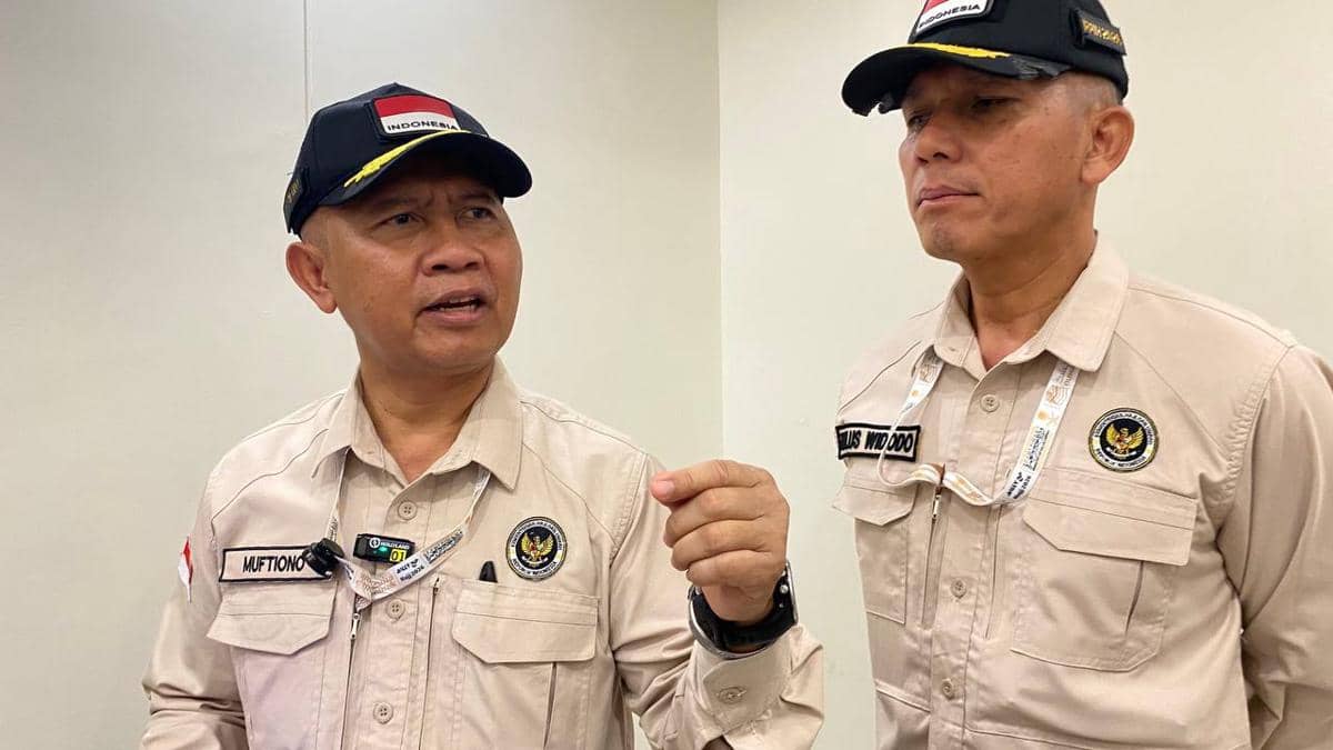 Dua petugas PPIH Arab Saudi mengenakan seragam krem dan topi hitam bertuliskan Indonesia sedang berbincang di dalam ruangan.