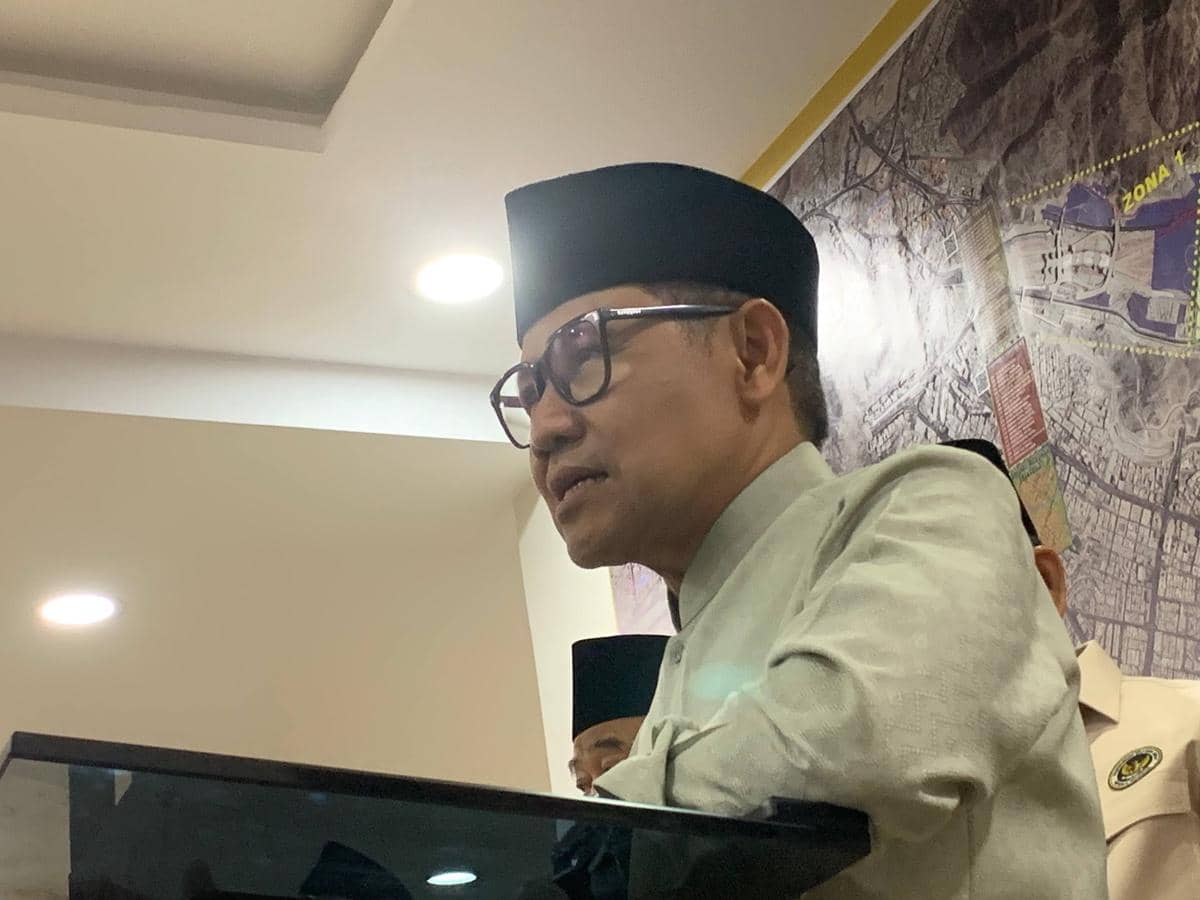 Seorang pejabat mengenakan peci hitam dan kemeja abu-abu berbicara di podium dengan latar peta besar di dinding belakang.