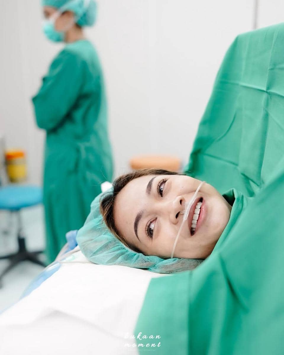 Seorang perempuan tersenyum di ruang operasi dengan mengenakan pakaian medis hijau dan selang oksigen di hidungnya.