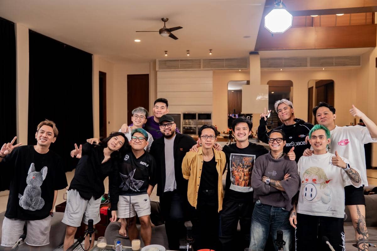 Sekelompok orang berpose bersama di dalam ruangan modern dengan pencahayaan hangat, tersenyum dan menunjukkan gestur damai ke kamera.