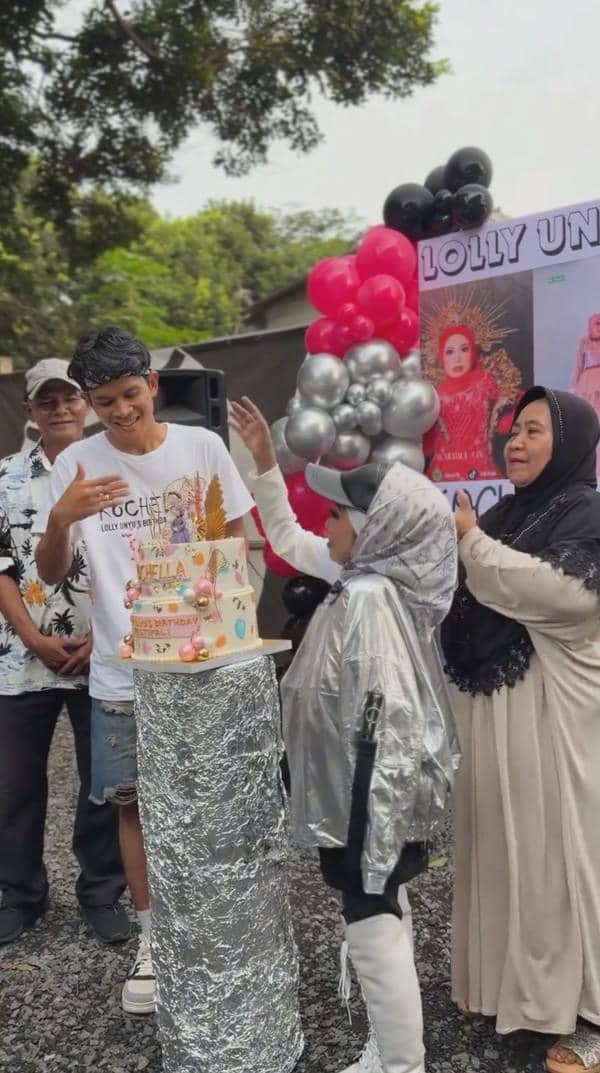 Beberapa orang merayakan ulang tahun dengan kue bertingkat dan balon berwarna merah, hitam, serta perak di luar ruangan.