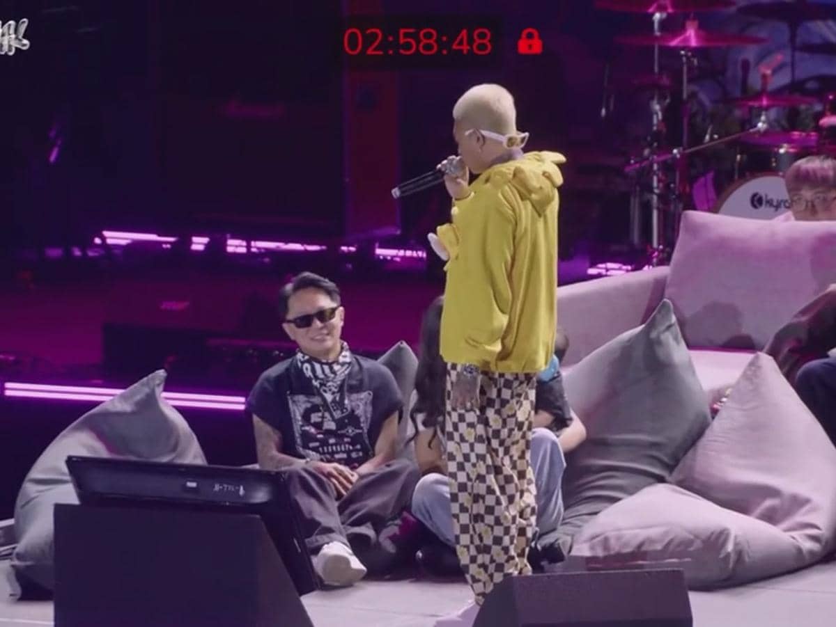 Seorang perempuan berjaket kuning berbicara di atas panggung dengan mikrofon di hadapan penonton yang duduk di sofa besar saat acara musik.
