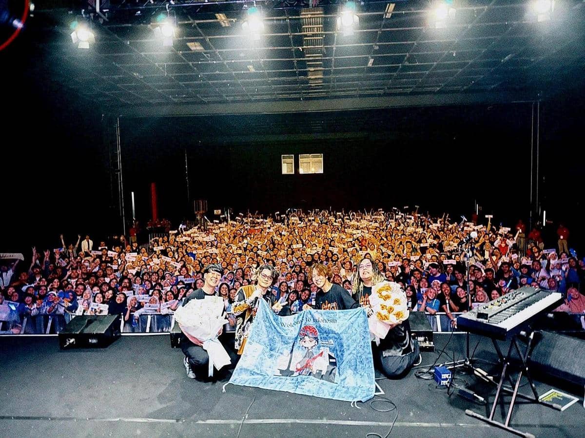 Para anggota band berpose di atas panggung bersama penonton yang memenuhi venue konser di Jakarta sambil memegang spanduk dan bunga.