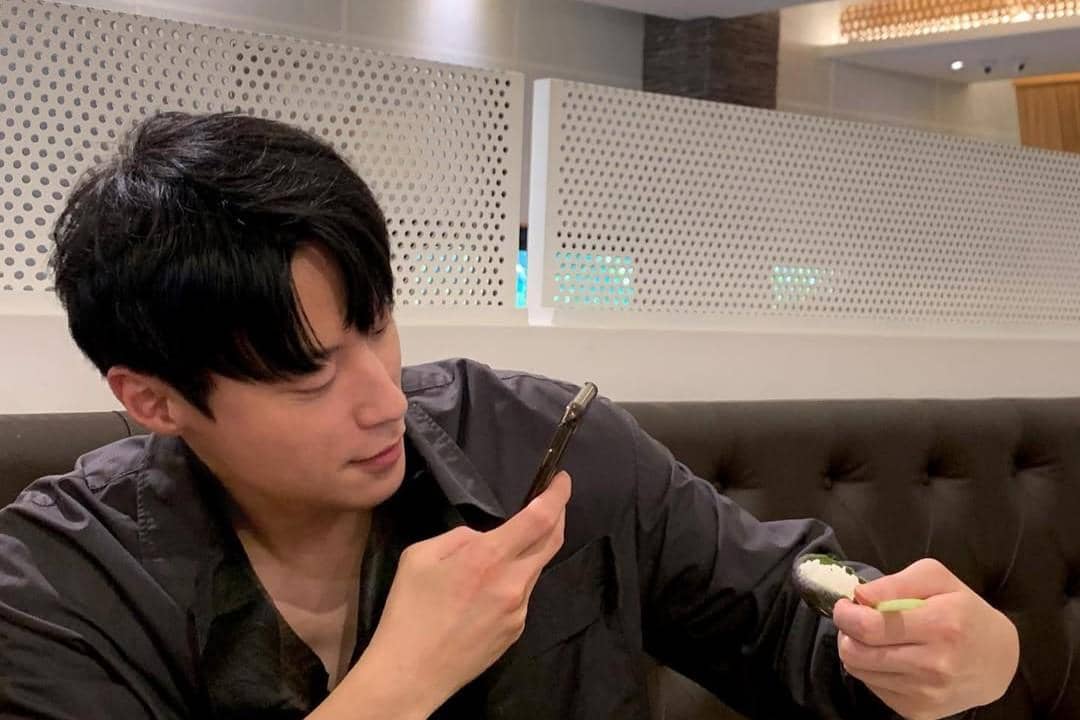 Seorang pria berambut hitam mengenakan kemeja gelap sedang memotret makanan dengan ponsel di dalam restoran modern.