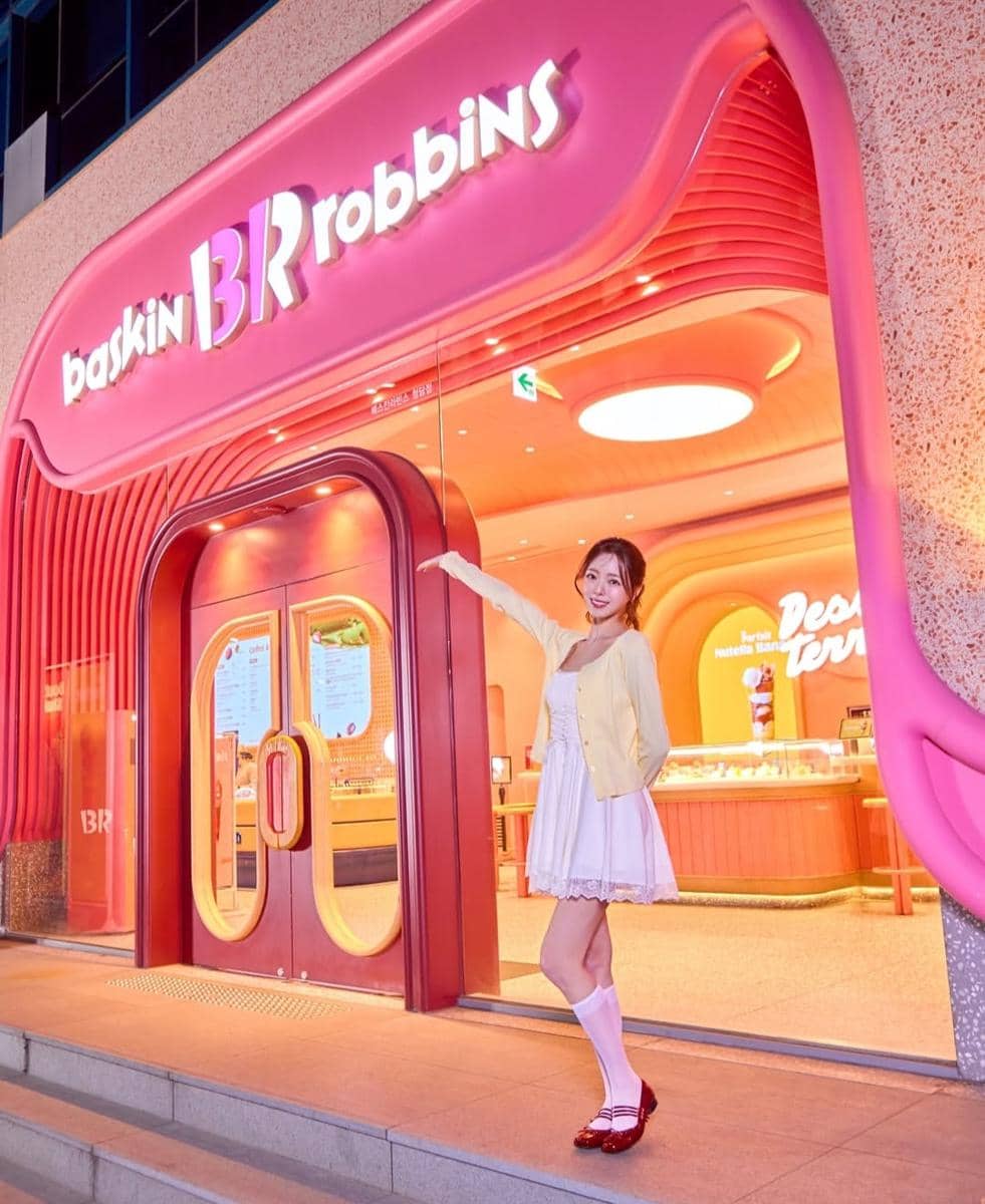 Seorang wanita mengenakan dress putih dan kardigan kuning berpose di depan toko es krim Baskin Robbins dengan pencahayaan berwarna pink cerah.
