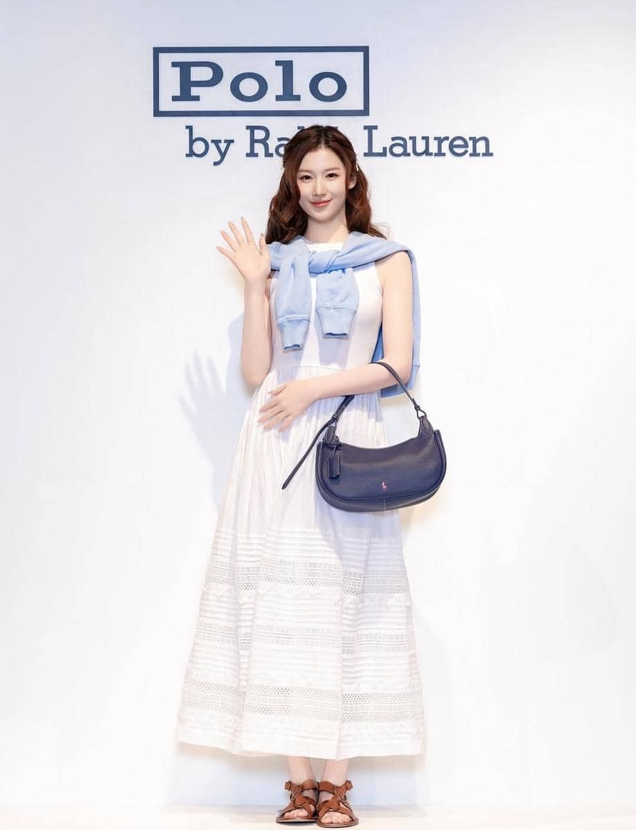 Seorang wanita mengenakan dress putih panjang dengan sweater biru muda di bahu dan tas bahu biru, berpose di depan logo Polo by Ralph Lauren.