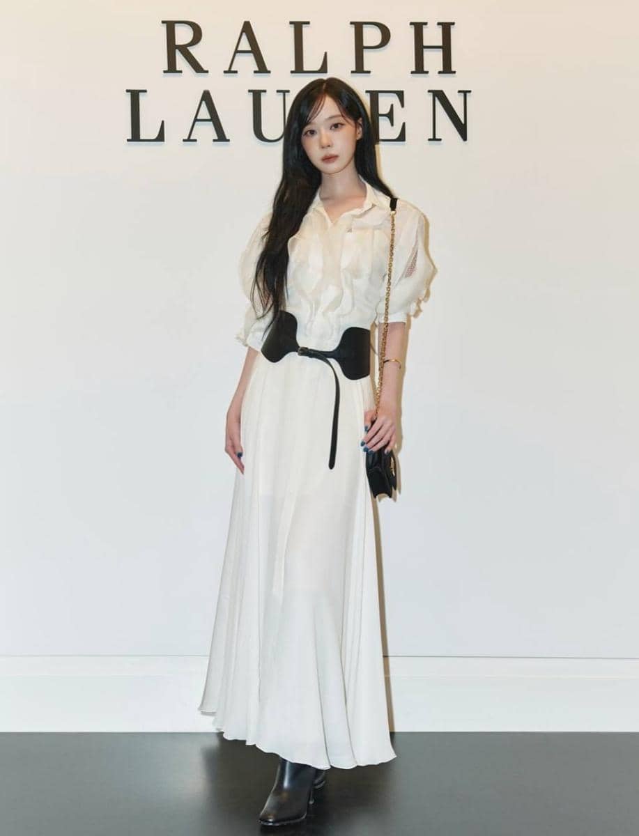 Seorang wanita berambut panjang mengenakan dress putih panjang dengan sabuk hitam dan tas rantai emas berdiri di depan logo Ralph Lauren.