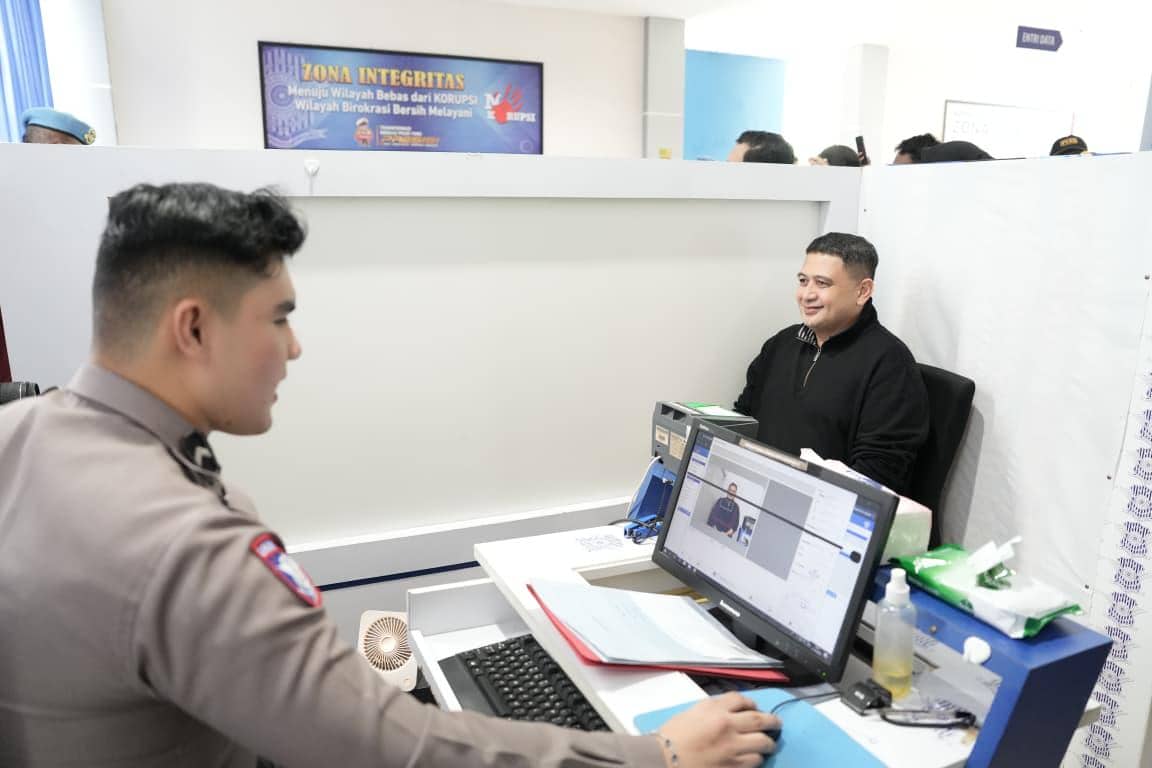 Seorang pria duduk tersenyum di depan petugas kepolisian saat proses pembuatan SIM C1 di ruang pelayanan dengan komputer dan peralatan administrasi.