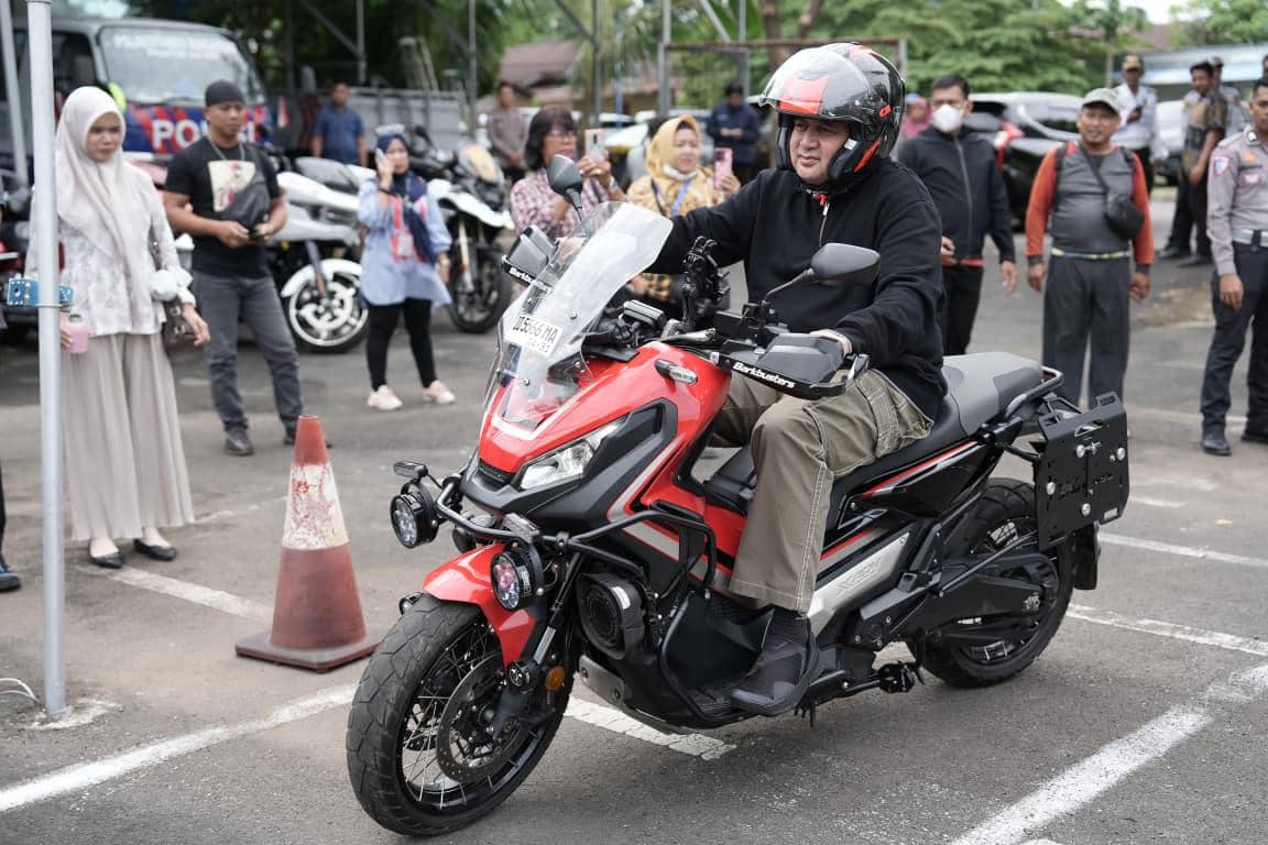 Seorang pria mengenakan helm dan jaket hitam mengendarai motor besar berwarna merah saat uji praktik SIM C1 di area parkir.