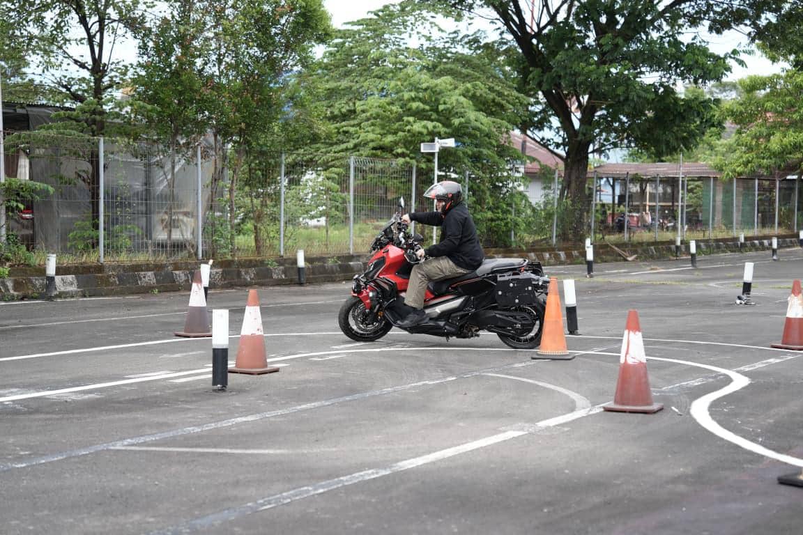 Seorang pengendara motor besar berjaket hitam dan helm sedang mengikuti uji praktik SIM C1 di area lintasan dengan rintangan cone.