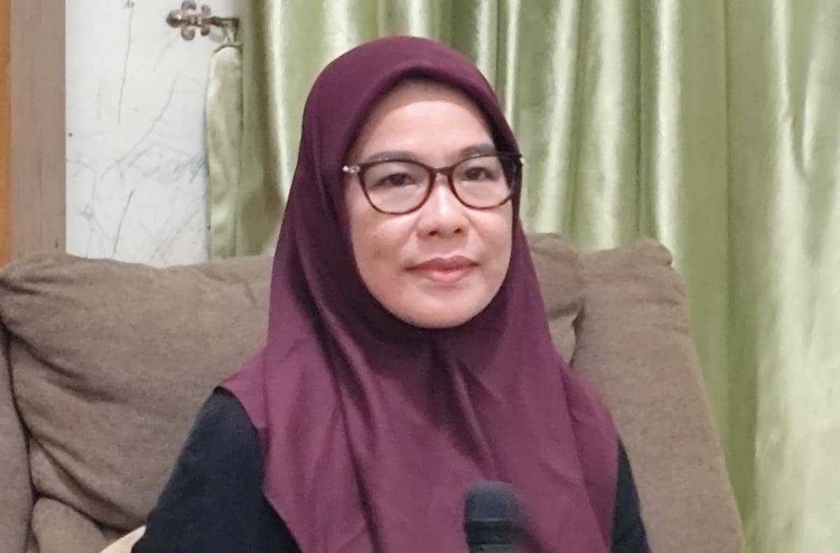 Seorang perempuan berhijab ungu tua dan berkacamata duduk di sofa sambil memegang mikrofon di ruangan dengan tirai hijau.
