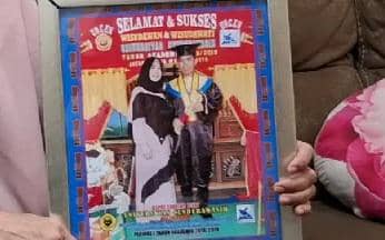 Seseorang memegang bingkai foto wisuda yang menampilkan dua orang mengenakan toga dan jubah wisuda di atas panggung.