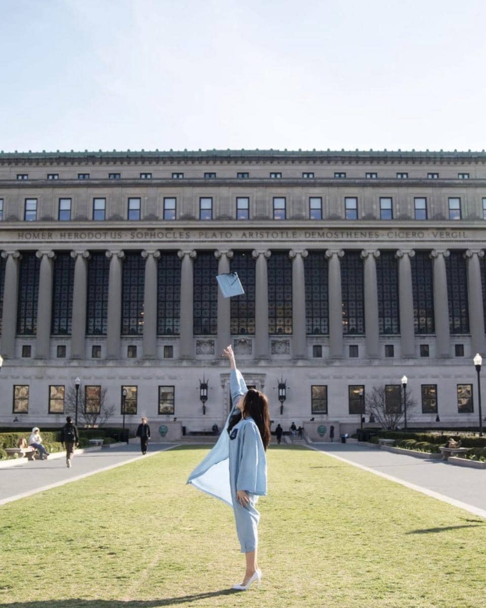 Seorang wisudawati mengenakan toga biru muda melempar topi wisuda di depan gedung kampus Columbia University.